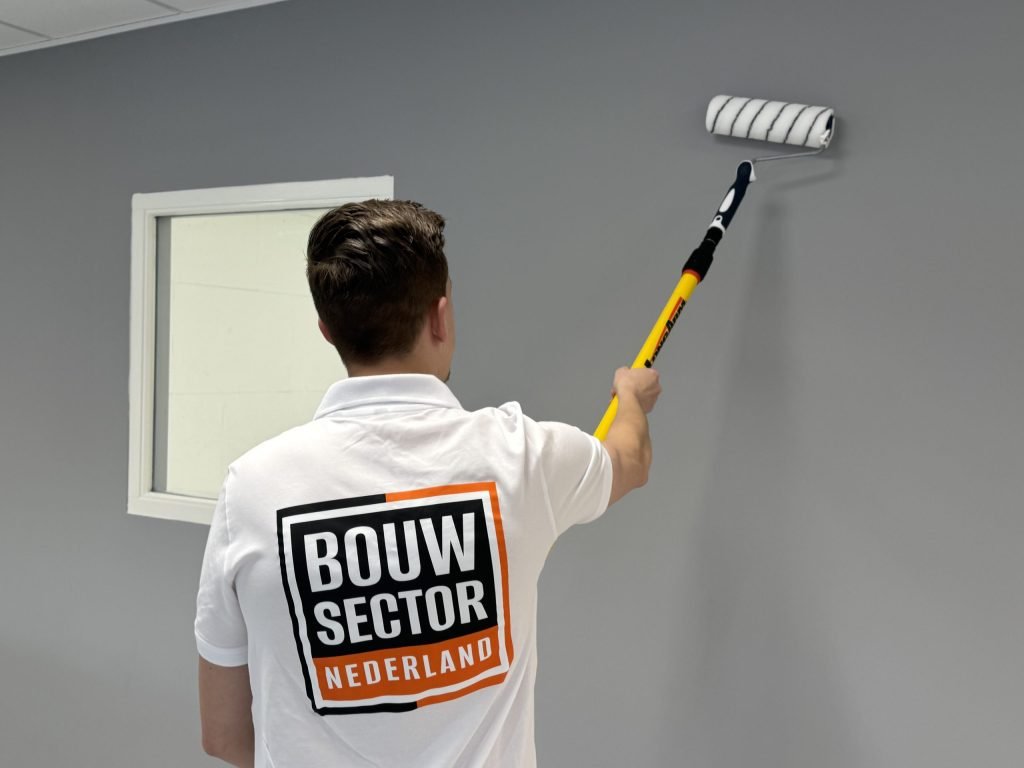 Schilder van Bouwsector Nederland rolt een grijze muur naast een raam met een roller op een verlengsteel