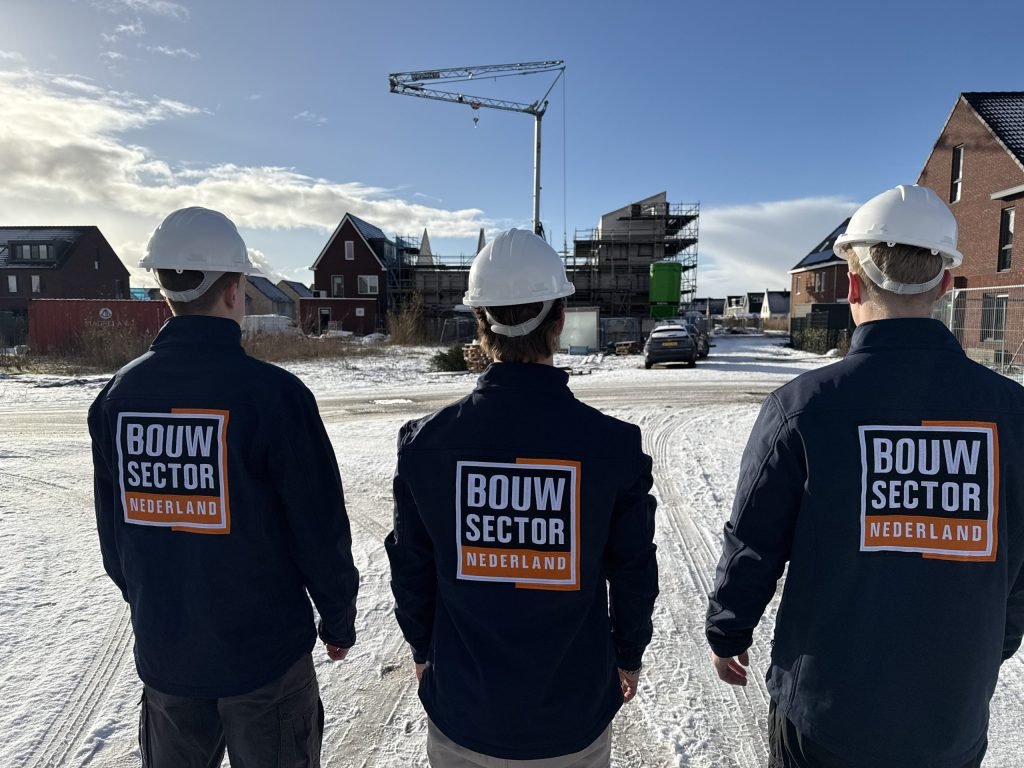 Drie bouwvakkers kijken naar een rij nieuwbouwwoningen in aanbouw onder een blauwe winterlucht